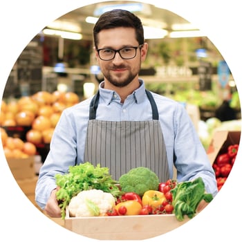 Mitarbeiter mit Brille und Schürze hält eine Kiste mit frischem Gemüse im Markt. Lächelt freundlich.